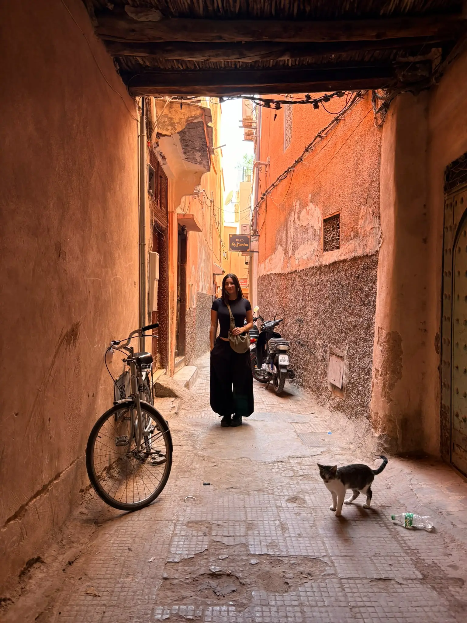 Amalia sonriendo en un callejón de la medina de Marrakech, con una bici apoyada en la pared y un gato en primer plano
